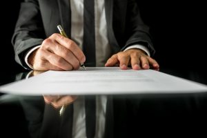 Front view of businessman signing important legal document on black desk with reflection.